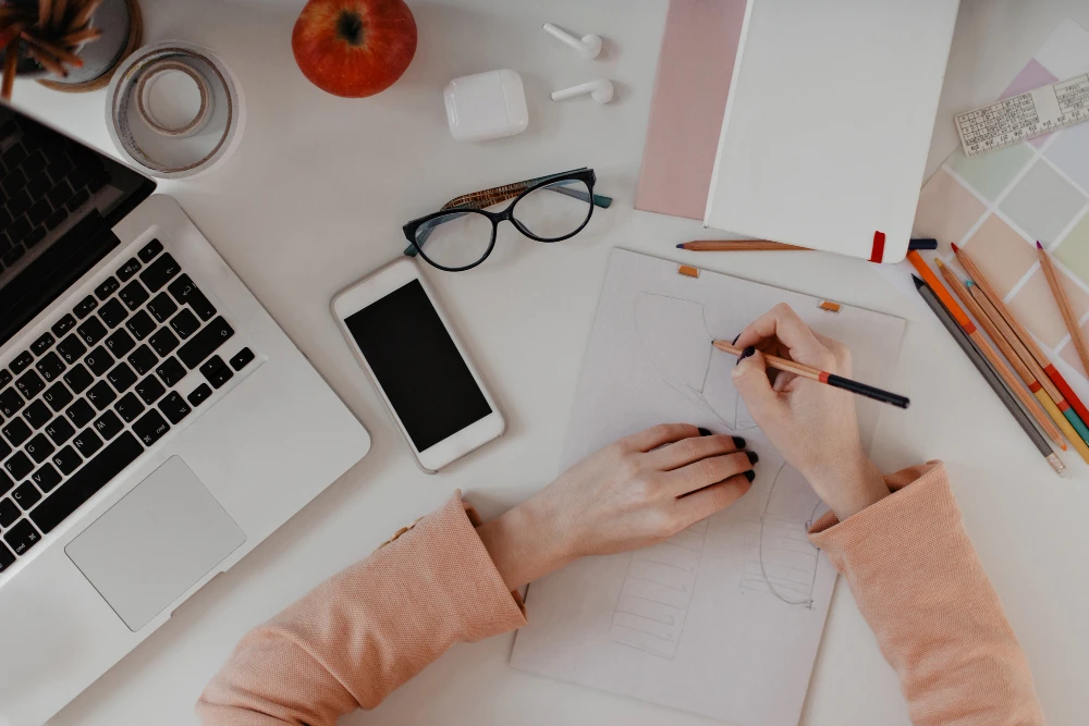 A person sketching a design concept at a desk with a laptop, smartphone, and stationery.