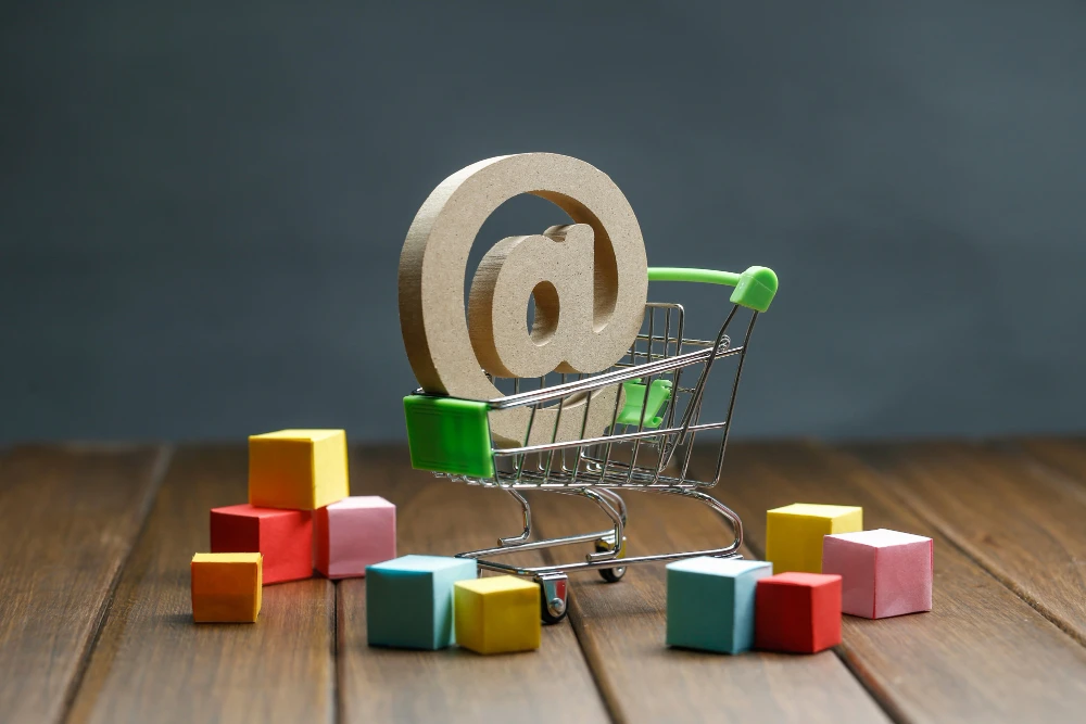 A miniature shopping cart with a wooden "@" symbol, surrounded by colorful blocks, representing online shopping.