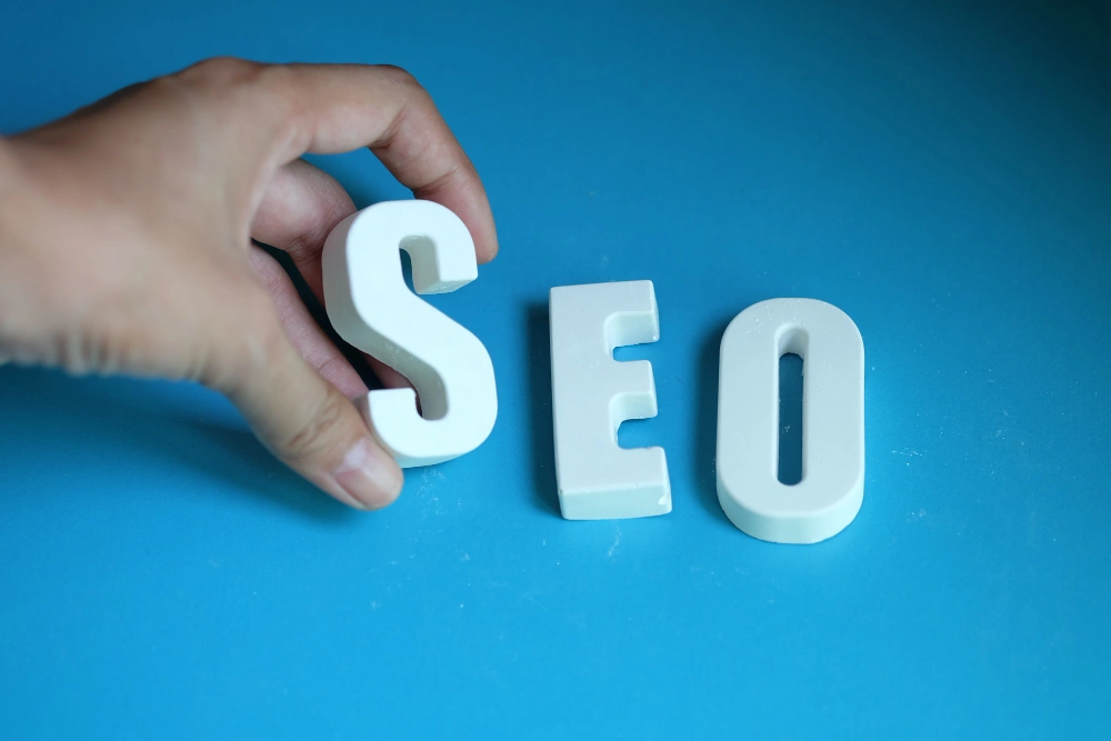 A hand placing the letter "S" to form "SEO" on a blue background, symbolizing search engine optimization.